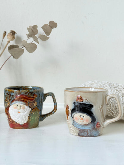 Pair of Christmas mugs with Santa and snowman designs photographed on a white surface.