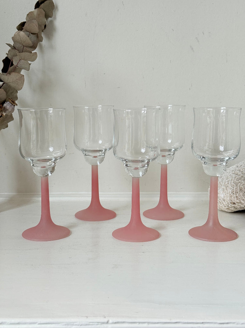 Set of five vintage wine glasses with clear bowls and pink stems photographed on a white surface.