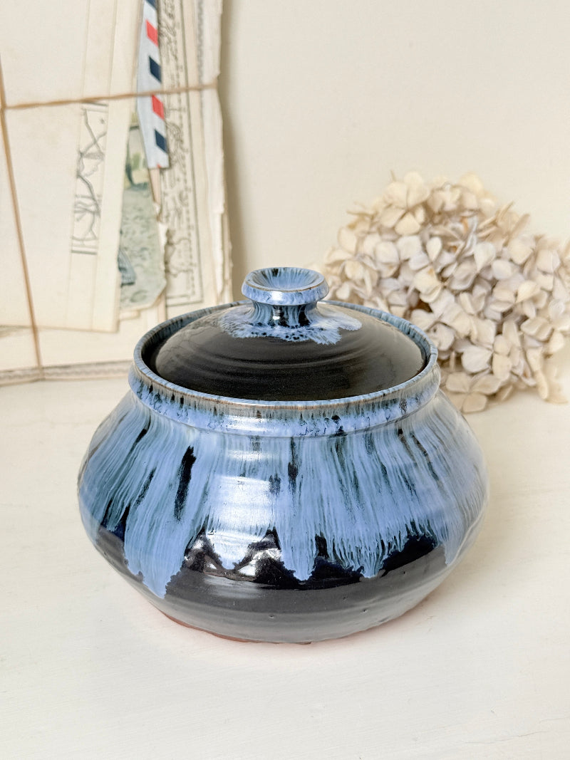 A round ceramic lidded jar with a glossy blue and black glaze, set against a neutral background with dried flowers and vintage papers.