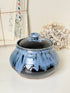 A round ceramic lidded jar with a glossy blue and black glaze, set against a neutral background with dried flowers and vintage papers.
