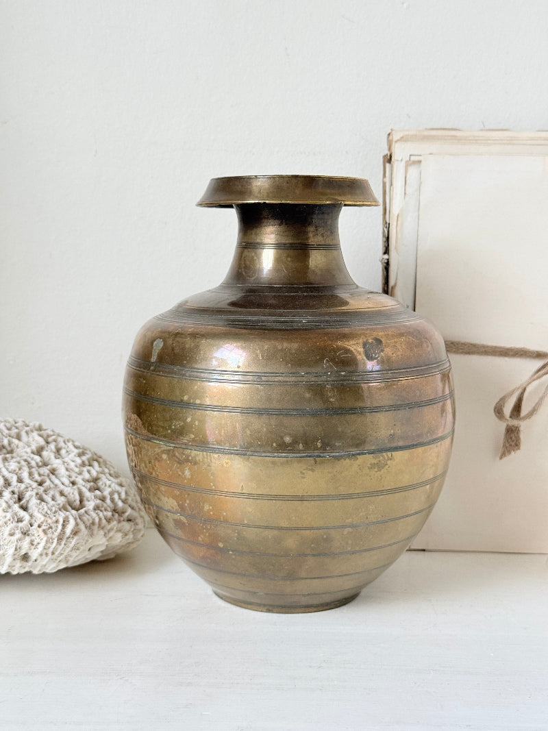 A vintage brass-colored metal vase with horizontal ridges, displayed on a white surface next to a white textured pillow and a white box.