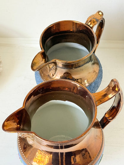 Two vintage copper pitchers with blue and white patterned bases, shown on a white surface against a white background.