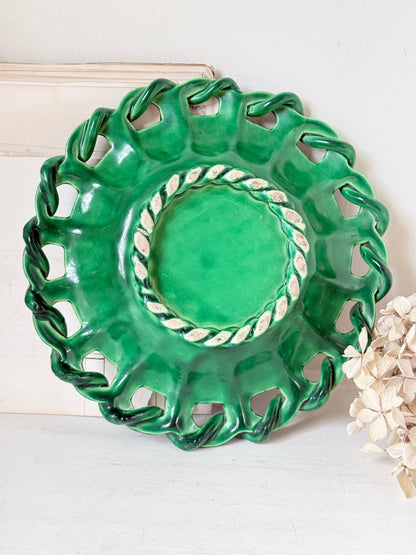 A green ceramic decorative plate with a scalloped, cutout rim and a twisted rope detail around the inner edge, displayed against a light background with dried flowers at the side.