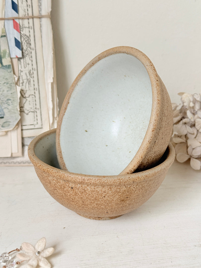 Two stacked ceramic bowls with a speckled beige exterior and a pale mint-green glazed interior, styled on a white tabletop with dried flowers and a vintage postcard frame in the background.