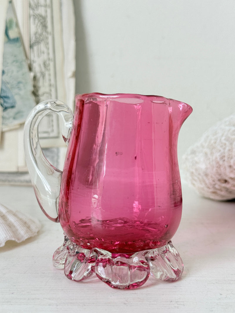 Antique cranberry glass cream jug with clear applied handle, crimped pedestal foot and deep pink hand blown glass body