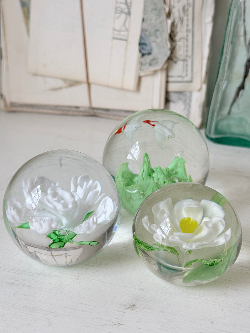 Trio of white flower glass paperweights in graduated sizes with green leaves, photographed on a white surface