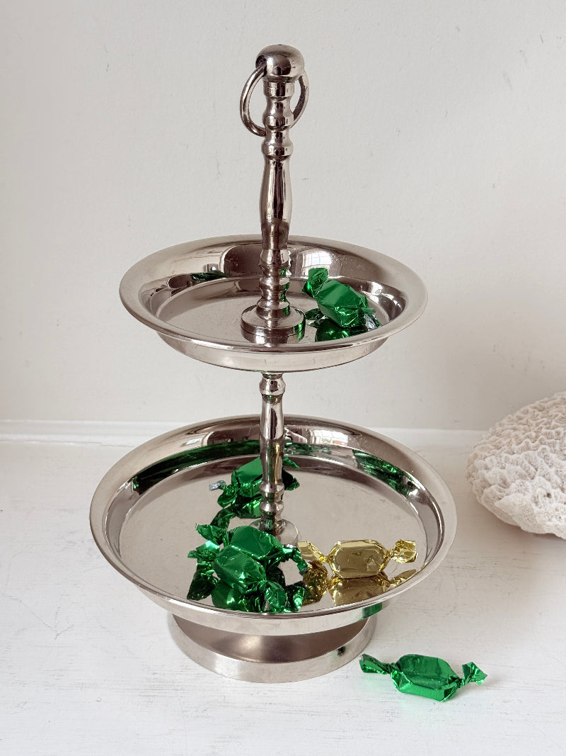 Two tier silver tone cake stand with polished reflective finish styled with green wrapped sweets on a white surface