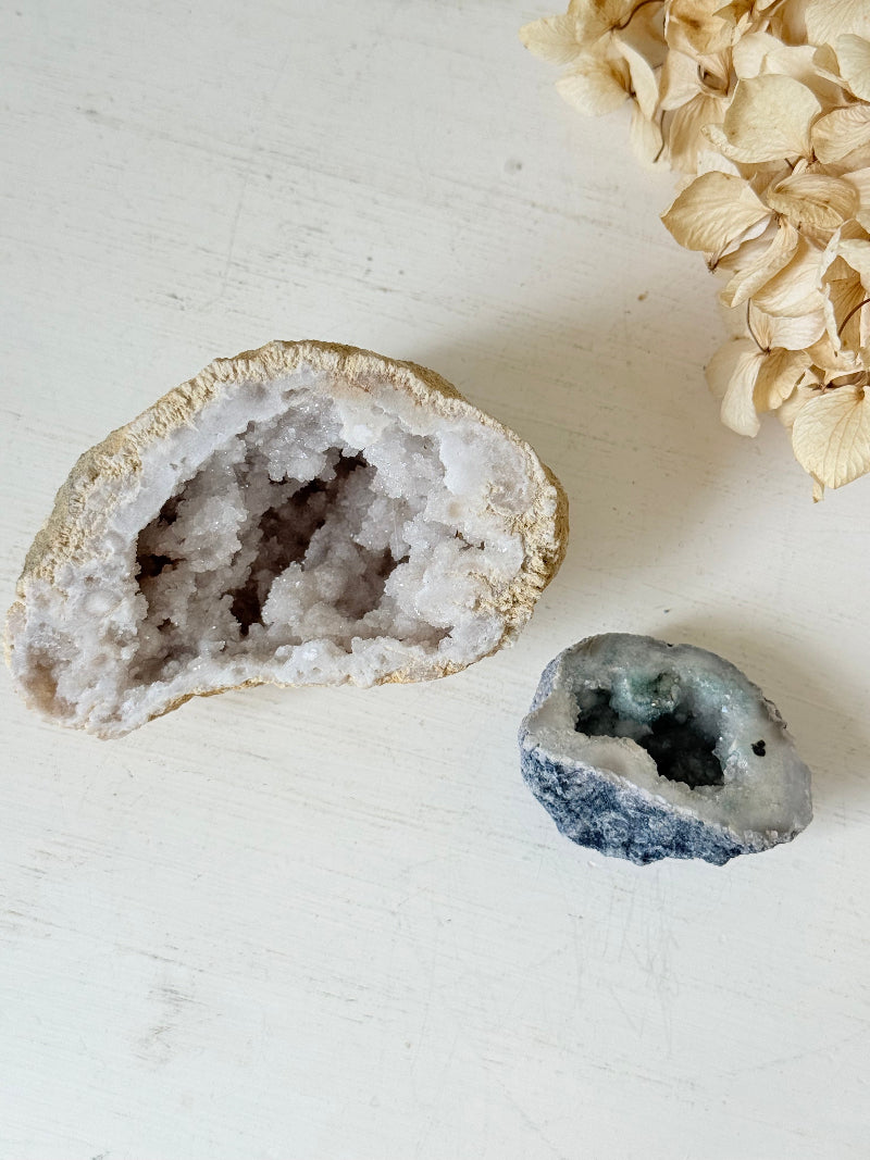 Pair of natural quartz geodes, one white and one lilac-toned, styled on a white surface with dried hydrangea