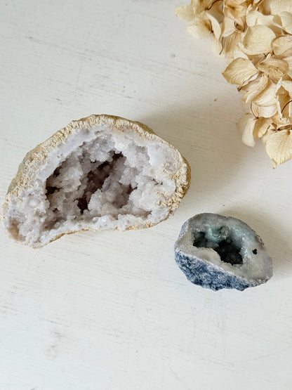 Pair of natural quartz geodes, one white and one lilac-toned, styled on a white surface with dried hydrangea