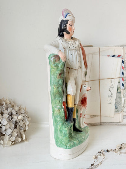 A vintage ceramic figurine of a man in historical military or hunting attire standing next to a green landscape panel and a dog, displayed on a white surface with decorative dried flowers and envelopes in the background.