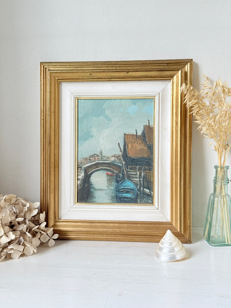 A framed painting of a canal scene with a bridge and boats, displayed on a white surface with decorative dried flowers and a glass vase against a white wall.
