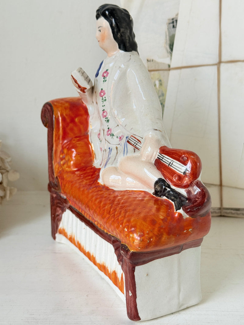 A vintage Chinese porcelain figurine depicting a man in a white floral jacket sitting on an orange-red patterned chaise lounge, holding a red instrument.