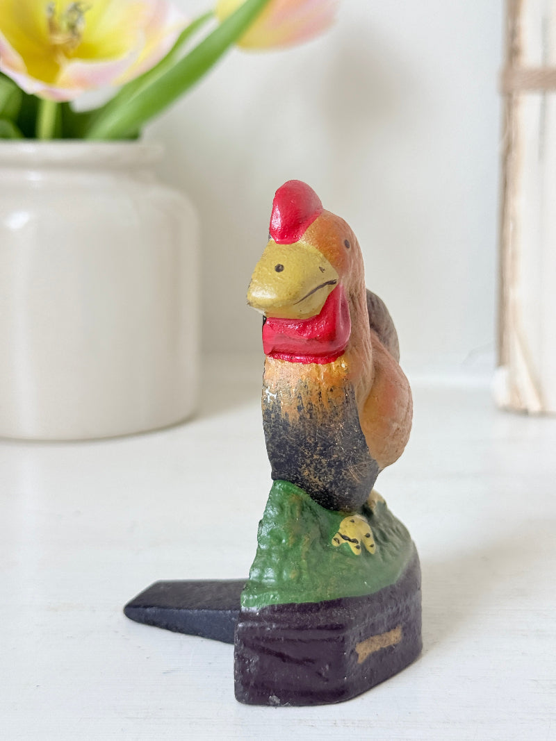 A small hand-painted wooden rooster figurine standing on a green hill with a black base, set against a softly blurred background with a white vase of tulips.