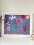A framed painting of three colorful flower pots against a textured purple and blue background, displayed on a white surface against a light beige wall.