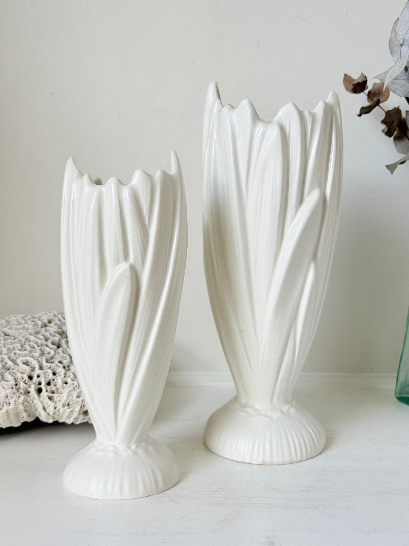 Two white ceramic tulip-shaped vases of different heights displayed on a light surface against a neutral off-white background.