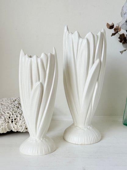 Two white ceramic tulip-shaped vases of different heights displayed on a light surface against a neutral off-white background.