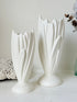 Two white ceramic tulip-shaped vases of different heights displayed on a light surface against a neutral off-white background.