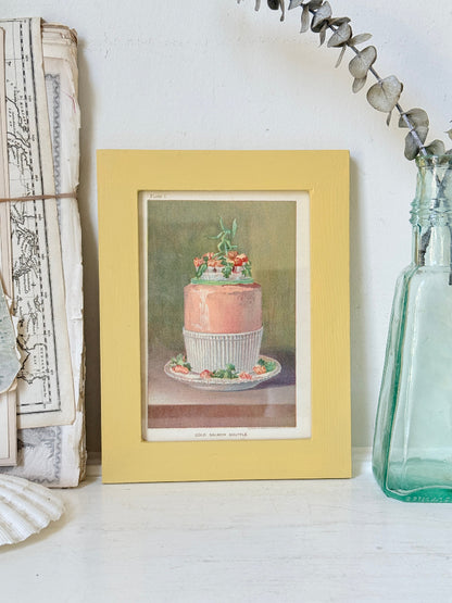 Framed Victorian cookery illustration of a celebration cake from 1896 in a mustard yellow frame displayed on a white surface.