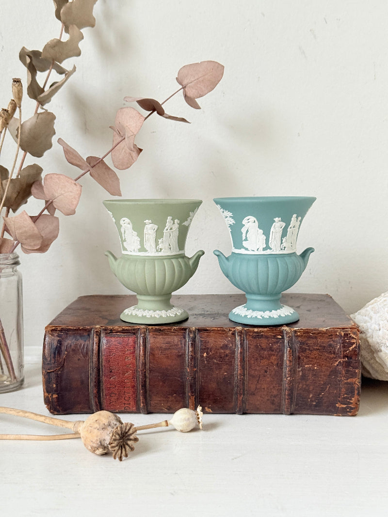 Pair of vintage Wedgwood Jasperware pedestal urns in sage green and pale blue, decorated with white neoclassical figures, styled on an antique leather book.