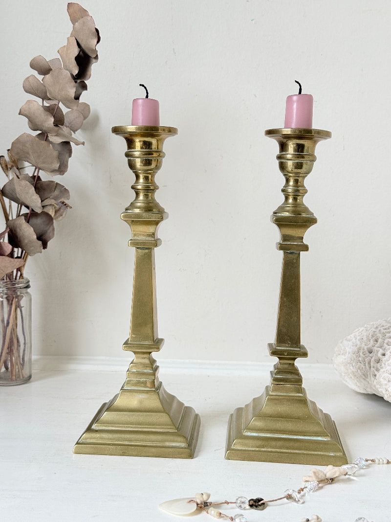 Pair of Georgian style brass candlesticks photographed on a white surface with tall tapered stems and stepped square bases holding pink taper candles