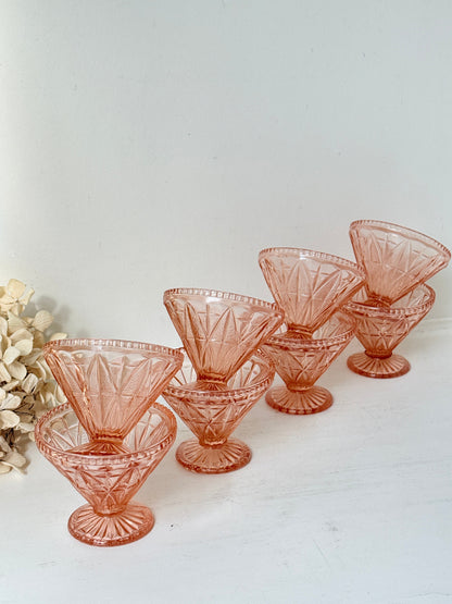 Six pink cut-glass cocktail glasses arranged on a white surface against a white background, with a small dried floral arrangement at the left edge.