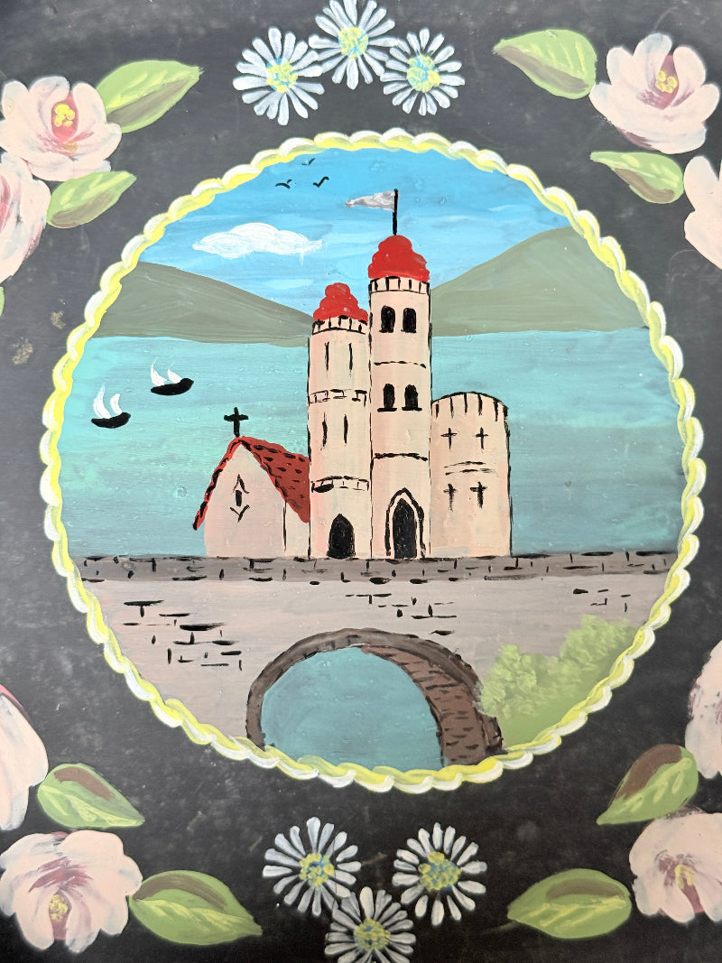 A hand-painted folk art print or panel featuring a castle and church on a stone wall by a blue lake, framed by a scalloped yellow border and surrounded by pink flowers and green leaves on a dark gray background.