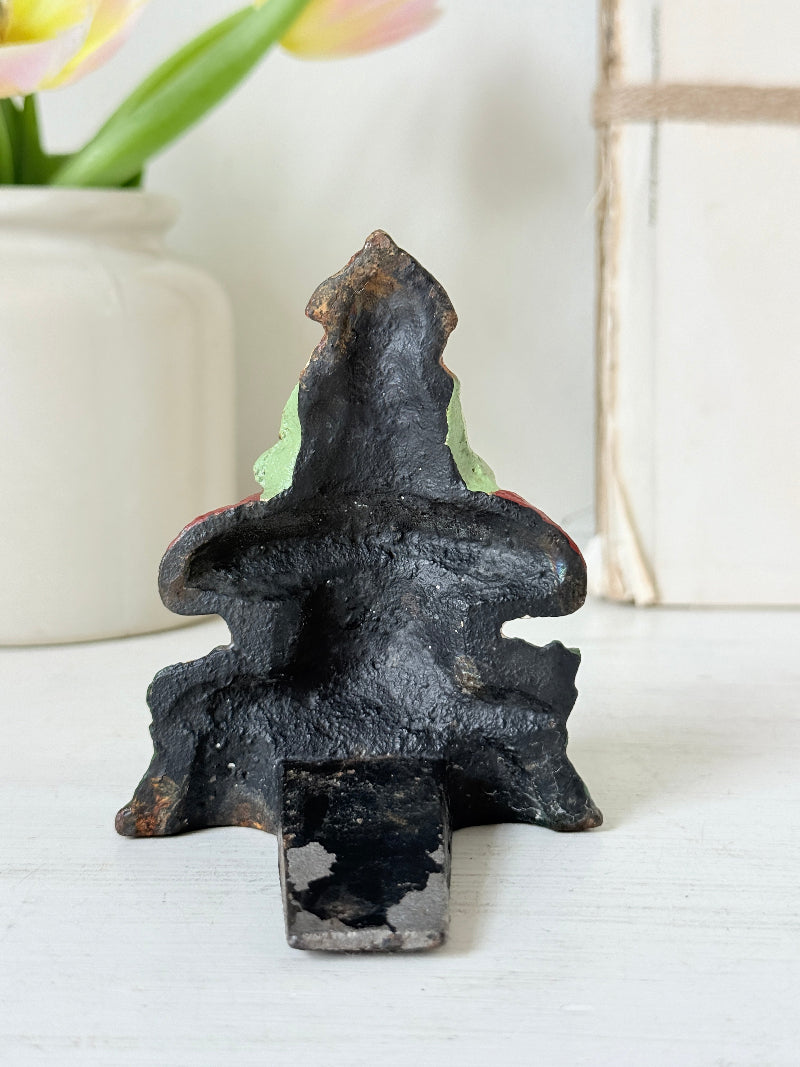 A rustic black cast iron fireplace hearth insert with a weathered, hand-painted design featuring light green and red accents, shown on a white surface.