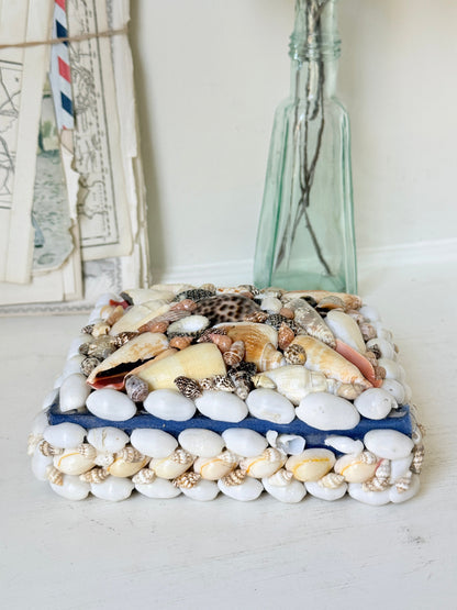 A rectangular decorative box or tray covered with white pebbles and assorted seashells, placed on a white tabletop with a tall translucent glass bottle behind it.