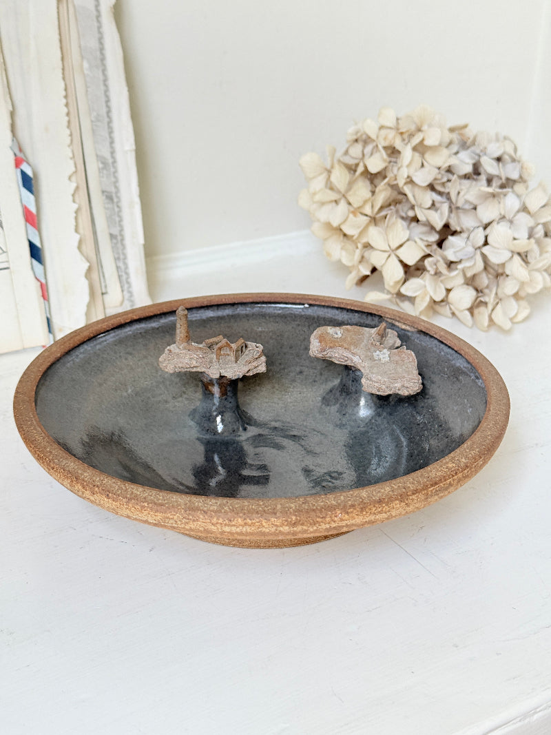 A round ceramic dish with a rustic brown rim and a dark gray glazed interior, featuring two small mounted natural specimens inside. The dish is styled on a white surface with dried flowers and books in the background.