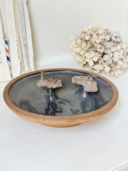 A round ceramic dish with a rustic brown rim and a dark gray glazed interior, featuring two small mounted natural specimens inside. The dish is styled on a white surface with dried flowers and books in the background.