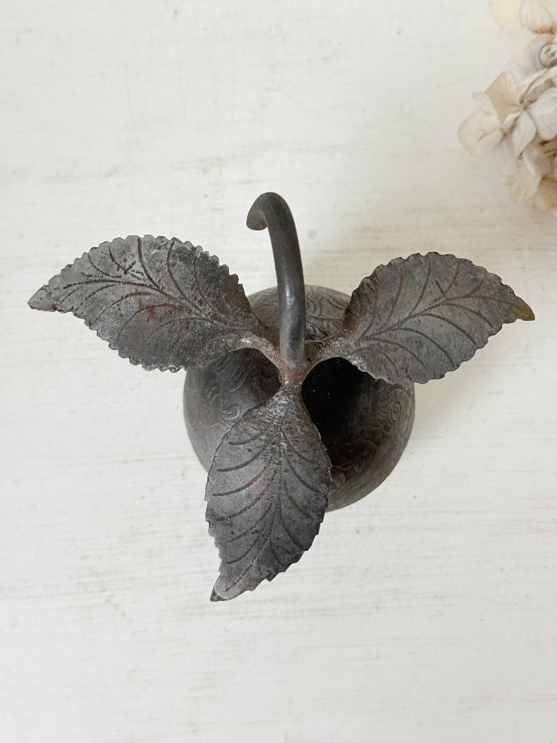 A dark metal decorative wall hook shaped like a curved stem with three leaf-like wings, photographed against a light off-white textured background.