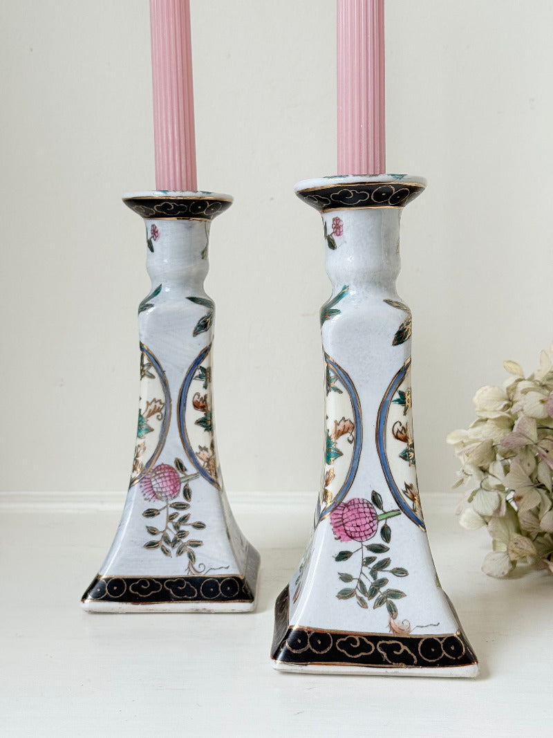 Pair of porcelain candle holders with hand-painted floral and vase motif in muted greens, pinks, and gold, styled with blush pink taper candles.