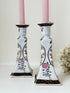 Pair of porcelain candle holders with hand-painted floral and vase motif in muted greens, pinks, and gold, styled with blush pink taper candles.
