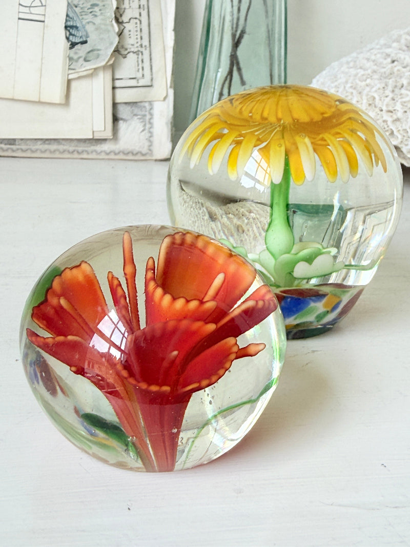 Pair of yellow and red flower glass paperweights with green stems, photographed on a white surface