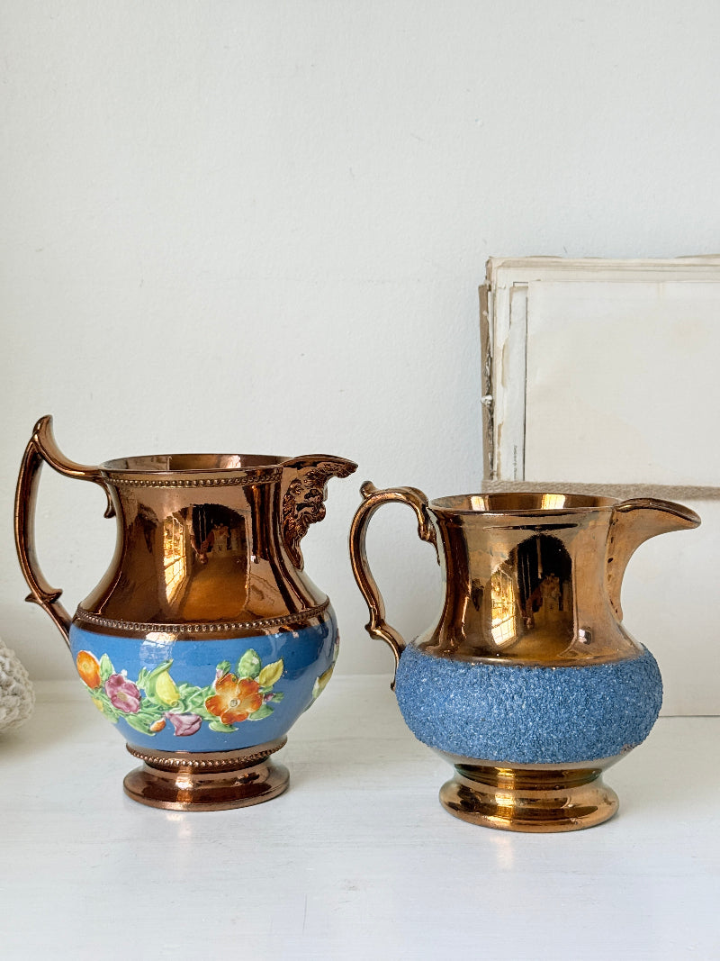 Two vintage-style ceramic pitchers with metallic copper-toned bodies and handles, one decorated with a blue floral band and the other with a textured blue lower section, displayed on a white surface against a white wall.