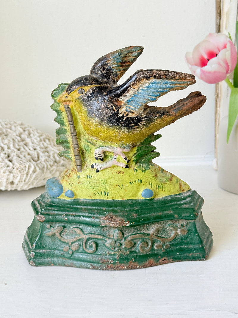 Vintage cast iron bird banknote holder shaped like a bird perched on a tree stump, with a green ornate base, photographed on a white surface with a pink tulip in the background.
