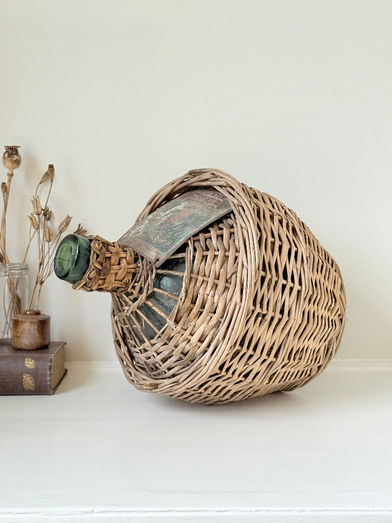 antique French demi john olive oil jar in wicker basket with green glass bottle and painted metal label, displayed on white surface
