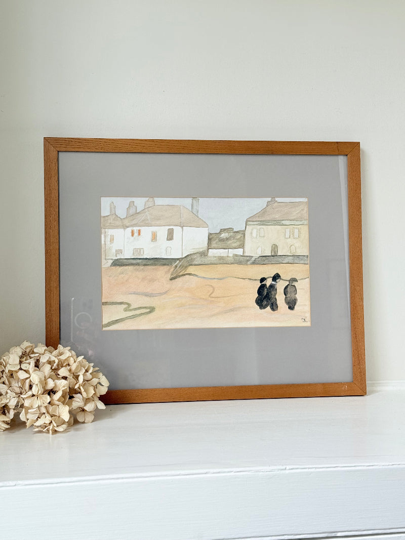 Framed neutral-toned village scene watercolour with figures in light oak-effect frame and pale grey mount, on white surface.