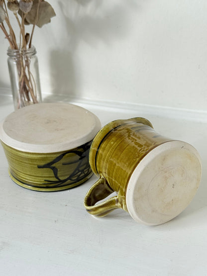 Studio pottery sugar bowl and milk jug in olive green glaze with black brushstroke decoration, photographed on a white surface.