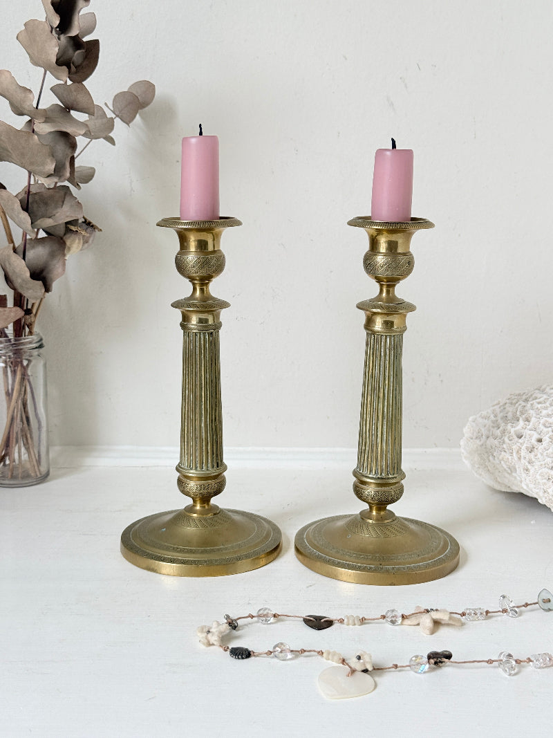 Pair of antique brass Empire style candlesticks photographed on a white surface with fluted columns and circular bases holding pink taper candles