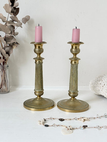 Pair of antique brass Empire style candlesticks photographed on a white surface with fluted columns and circular bases holding pink taper candles