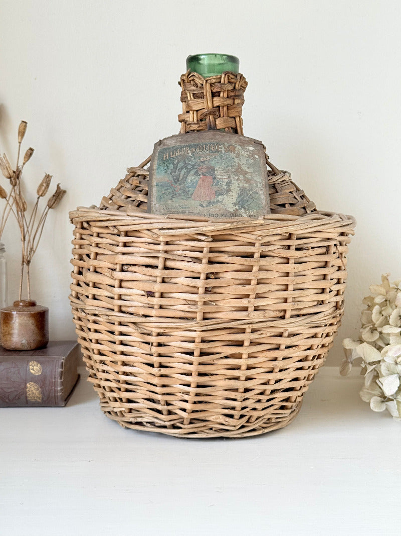 antique French demi john olive oil jar in wicker basket with green glass bottle and painted metal label, displayed on white surface