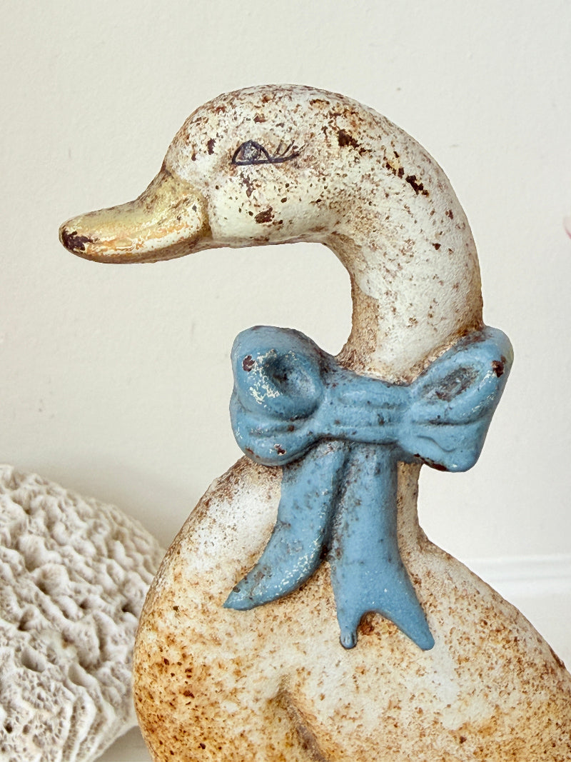 A decorative duck figurine with a speckled off-white body, a blue bow around its neck, and a gold-toned beak, shown against a light neutral background.
