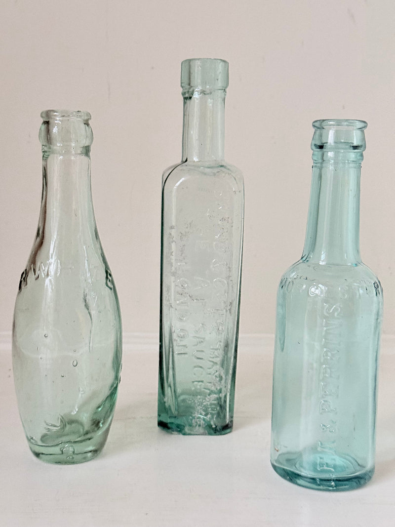 Trio of antique aqua glass bottles with embossed lettering, photographed on a white surface with dried stems