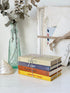 A stack of four vintage hardcover books tied with twine sits on a white surface next to a glass bottle with dried flowers and a shell. The book spines read &