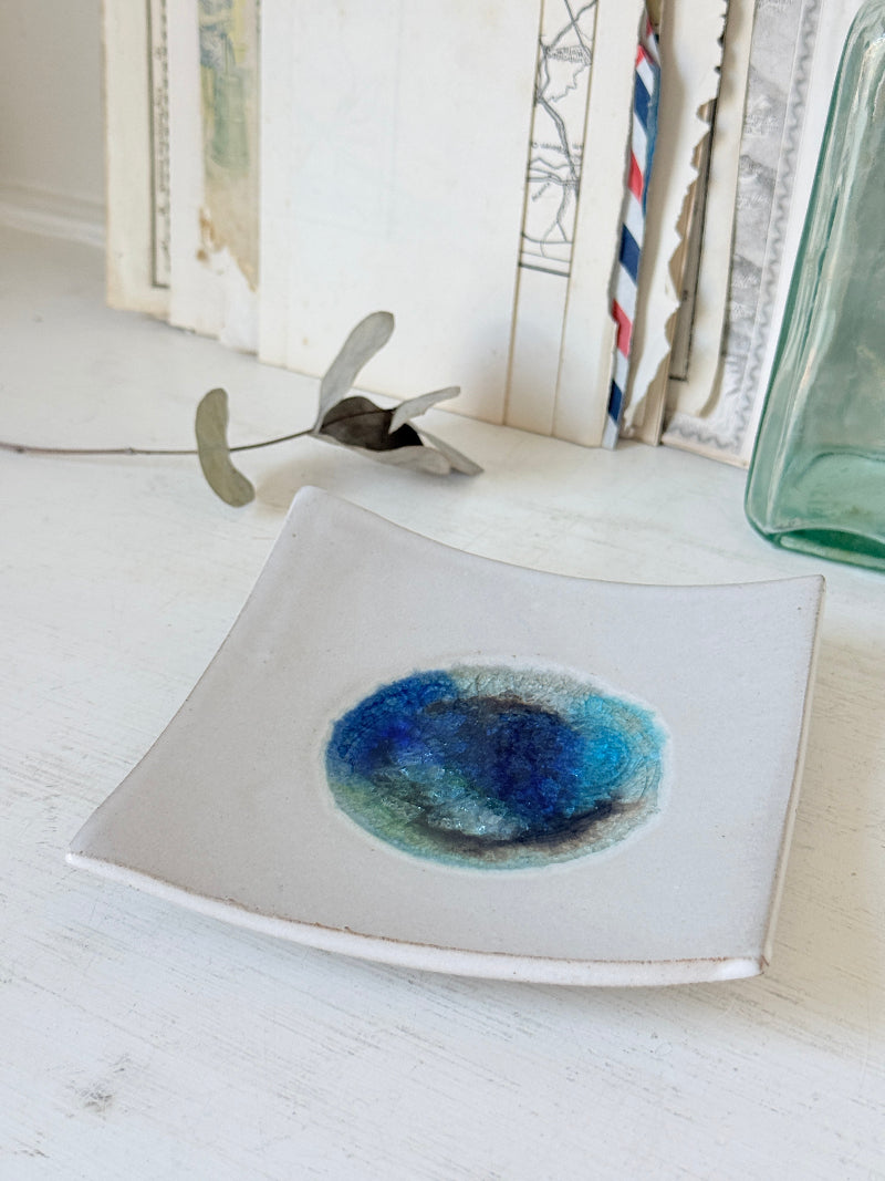 A square ceramic dish with a curved rim, featuring a hand-painted blue and green watercolor design in the center, styled on a white wooden surface with vintage books and a glass bottle in the background.