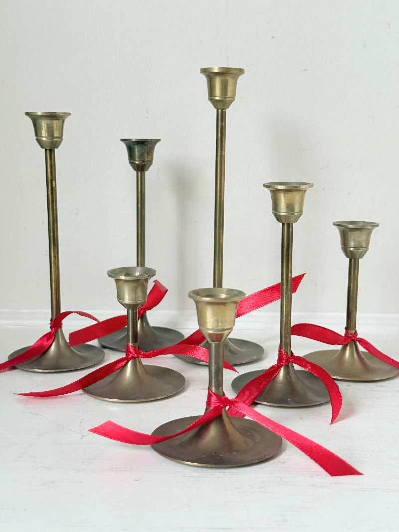 Set of 7 vintage brass candlesticks in graduated heights with aged brass finish styled with red ribbon on white surface
