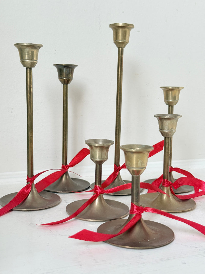 Set of 7 vintage brass candlesticks in graduated heights with aged brass finish styled with red ribbon on white surface
