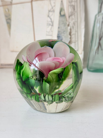 Large clear glass paperweight with a pink flower and green leaves inside, photographed on a white surface