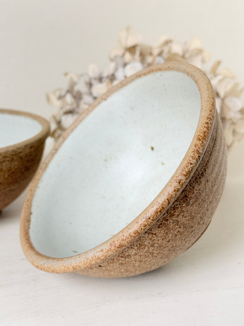 A close-up of a handmade ceramic bowl with a speckled brown exterior and a glossy pale blue-gray interior, shown on a light neutral surface with dried flowers in the background.